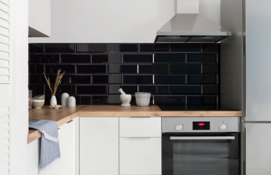 siyah parlak tezgah arasiduvar seramigi, zeminde cini motifli yer karosu, beyaz mutfak dolaplari ve ahsap tezgah ile kucuk ve acik plan mutfak