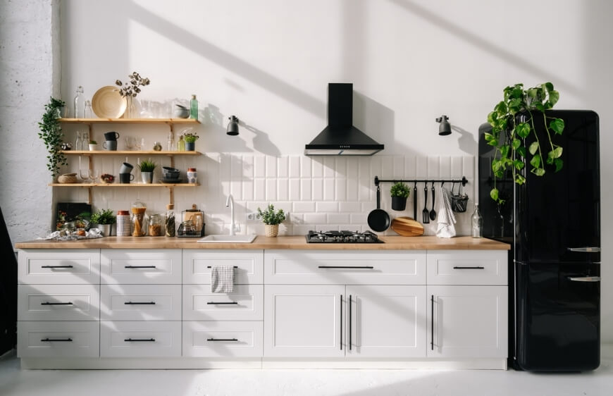 beyaz mutfak dolabi, ahsap mutfak tezgahi,siyah buzdolabi, acik ahsap duvar raflari, parlak ve ferah modern mutfakta beyaz tezgah arasi seramik