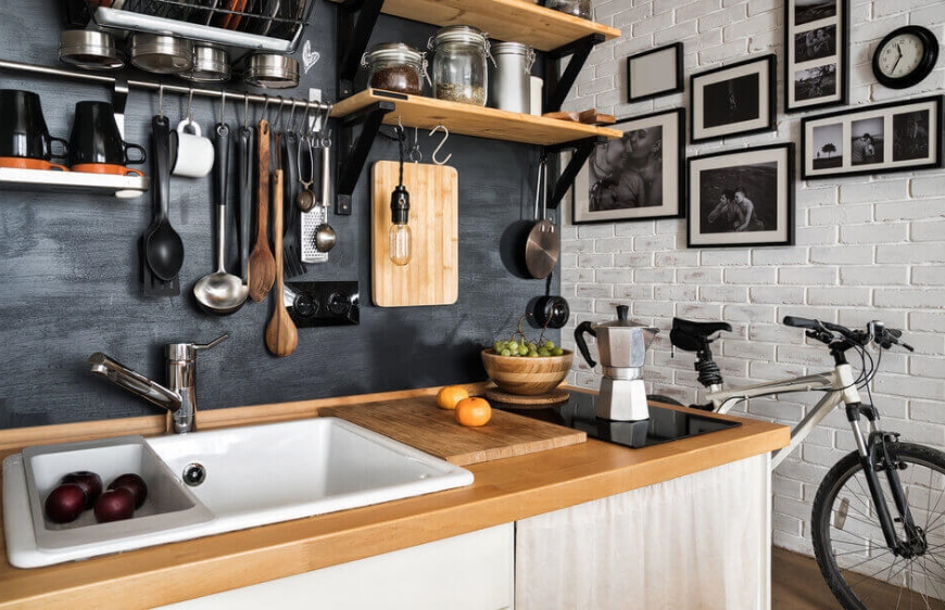 ahsap tezgahli mutfakta siyah tazgah arasi duvar boyasi ve duvara asilan mutfak gerecleri ve siyah beyaz tablolar ile kalabalik rustik mutfak stili