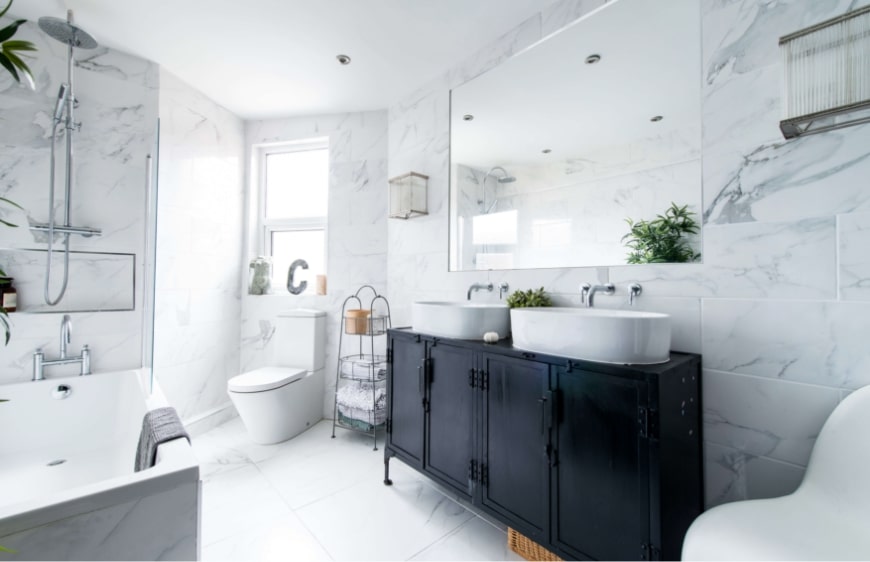 beyaz renklerin hakim oldugu cok koseli banyoda siyah endustriyel metal etajer, lavabo tezgah alti dolabi uzerinde ikiz lavabo ve ankastre krom armatur, pencere onunde klozet 